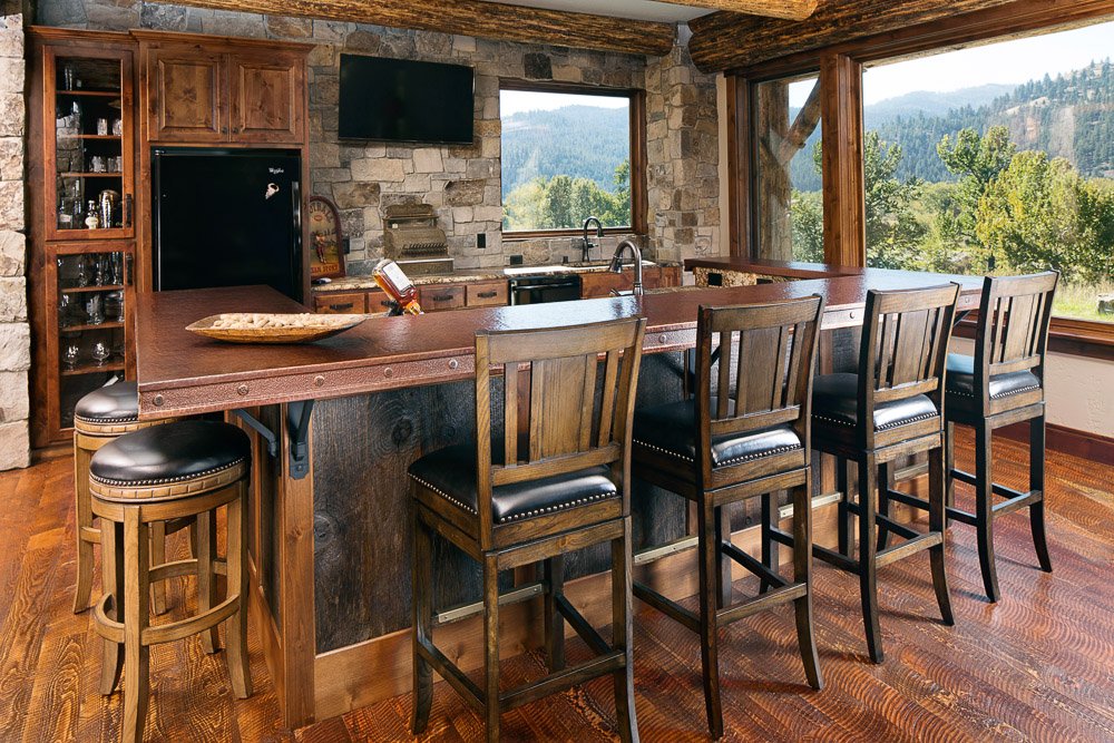 A rustic kitchen with a wooden island and four black leather stool chairs, stone walls, large windows showing a mountain landscape, and wooden beams overhead. Custom Home built in Montana by Edgell Building.