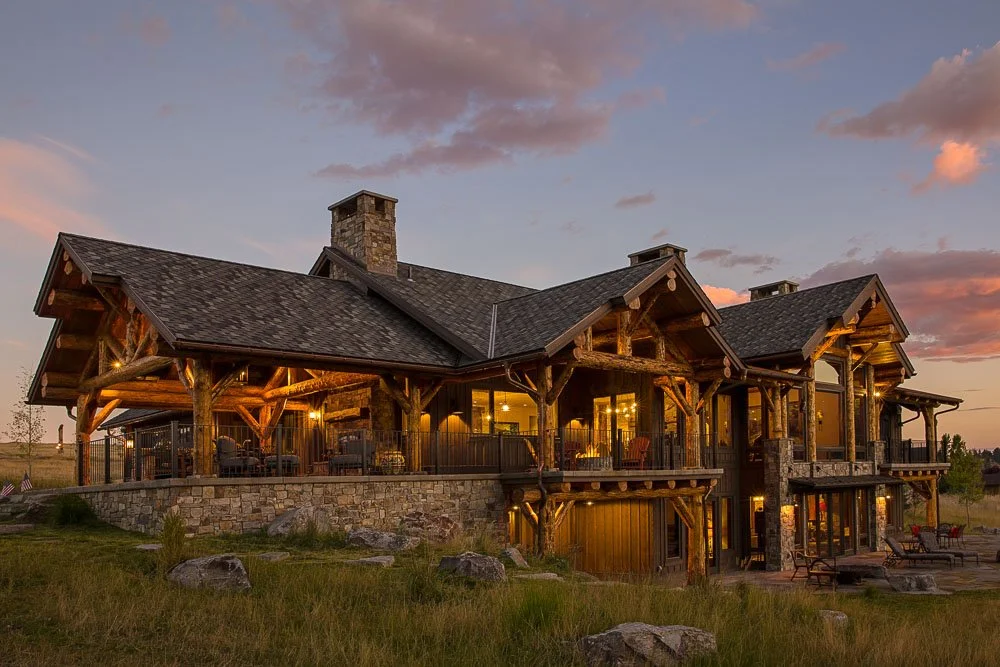A large house built with wood and stone, featuring a spacious porch with outdoor furniture, set on a grassy landscape during sunset. Custom Home built in Montana by Edgell Building.