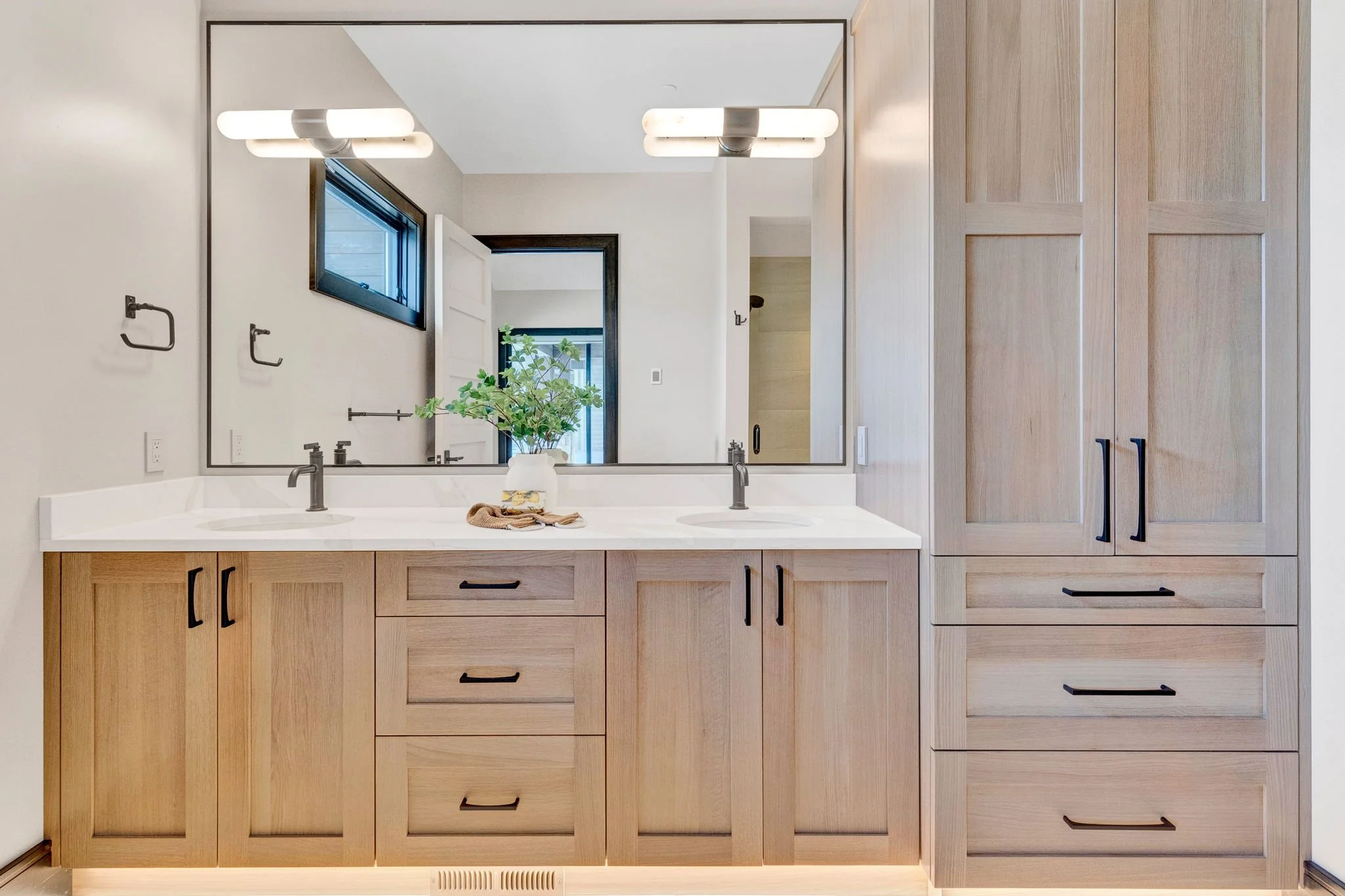 Bathroom vanity with dual sinks, large mirror, wooden cabinetry, black handles, and a potted plant on the countertop. Custom Home built in Montana by Edgell Building.