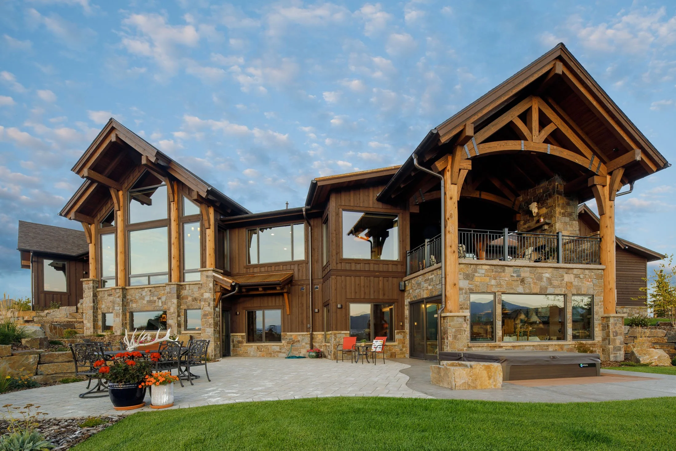 A large modern house with a rustic design featuring wooden beams, stone accents, multiple large windows, and a spacious outdoor patio with chairs, potted plants, and a hot tub. Custom Home built in Montana by Edgell Building.