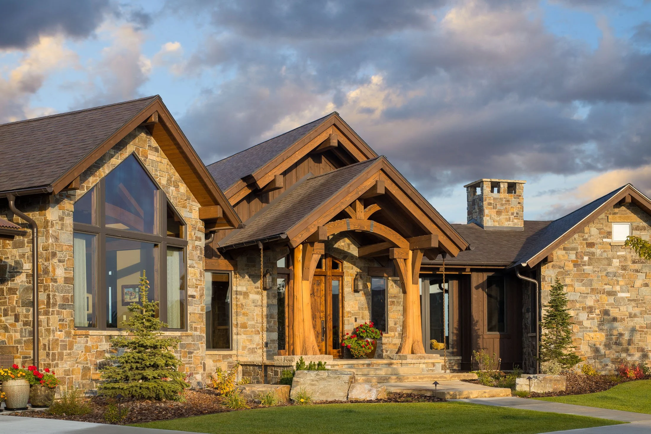 A house made of stone and wood with a front porch, surrounded by a well-maintained lawn and landscaping, under a partly cloudy sky. Custom Home built in Montana by Edgell Building.