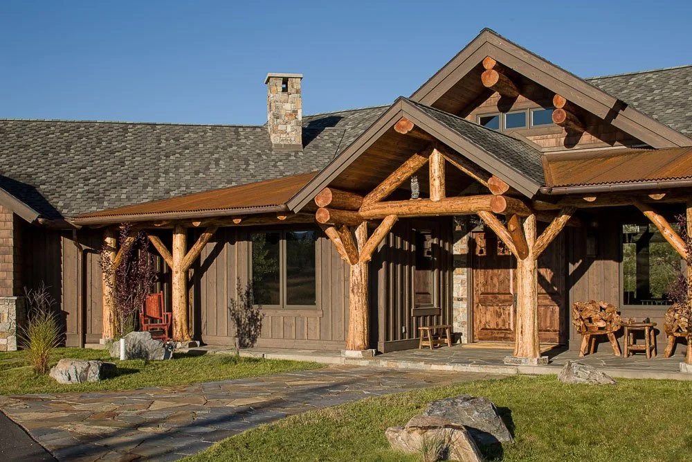 Front view of a rustic log cabin-style house with wooden support beams, stone chimney, stone walkway, and a small lawn with rocks. Custom Home built in Montana by Edgell Building.