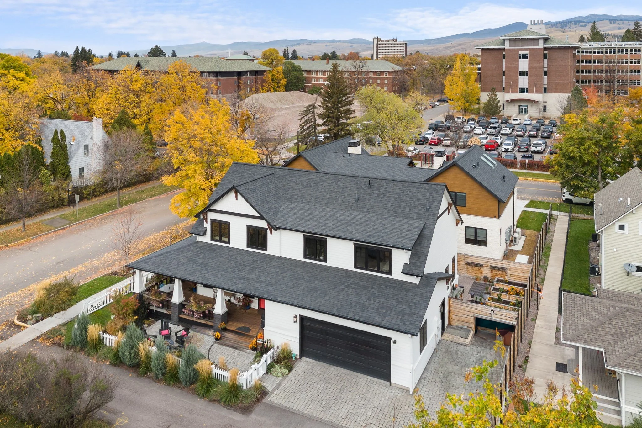 A large white and black two-story house with a front porch and a attached garage, surrounded by landscaped yard with trees and potted plants, located in a neighborhood with fall foliage. Custom Home built in Montana by Edgell Building.