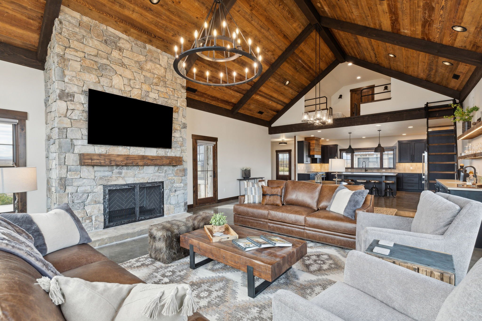 Living room with a stone fireplace, mounted flat-screen TV, wooden ceiling, brown leather and gray fabric sofas, coffee table, and modern open kitchen in the background. Custom Home built in Montana by Edgell Building.