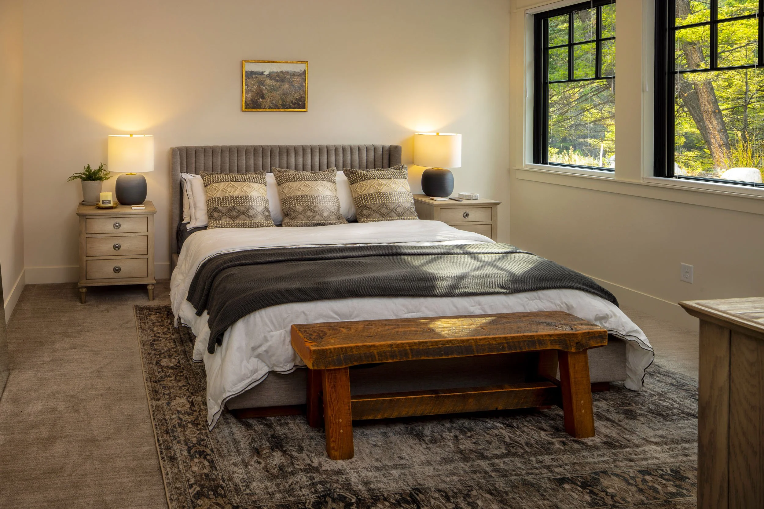 A cozy bedroom with a large bed, decorative pillows, nightstands with lamps, a painting, a wooden bench at the foot of the bed, and window views of lush greenery outside.