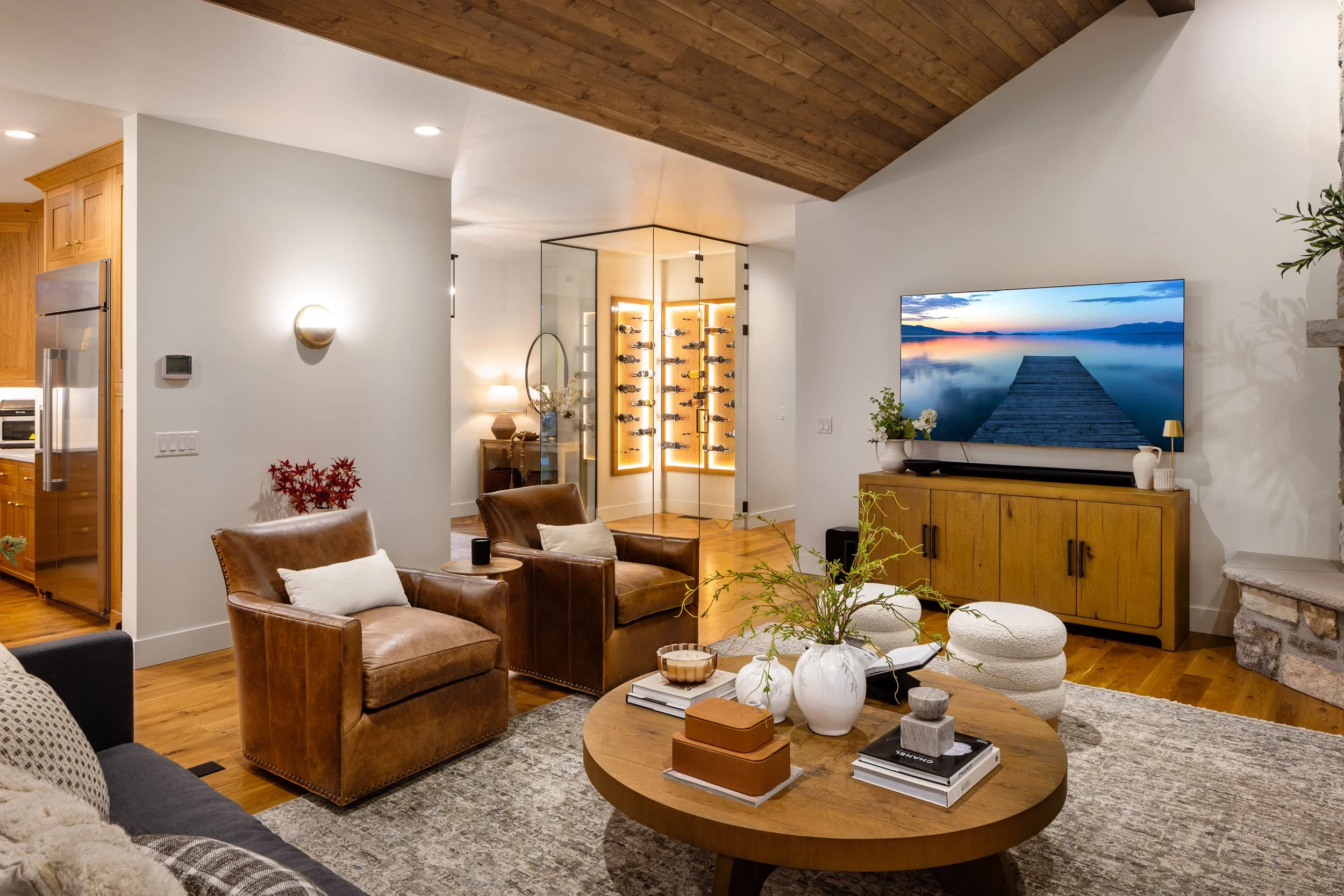 Living room with leather armchairs, a wooden coffee table with decorative vases and books, a large flat-screen TV on a wooden cabinet, a stone fireplace, and a staircase leading to a wine rack display area.