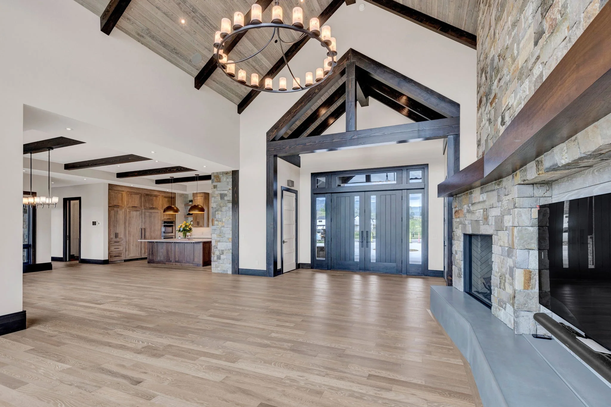 Spacious modern living room with wooden floors, a large stone fireplace, and a ceiling chandelier, featuring exposed wooden beams and large blue double doors leading outside. Custom Home built in Montana by Edgell Building.
