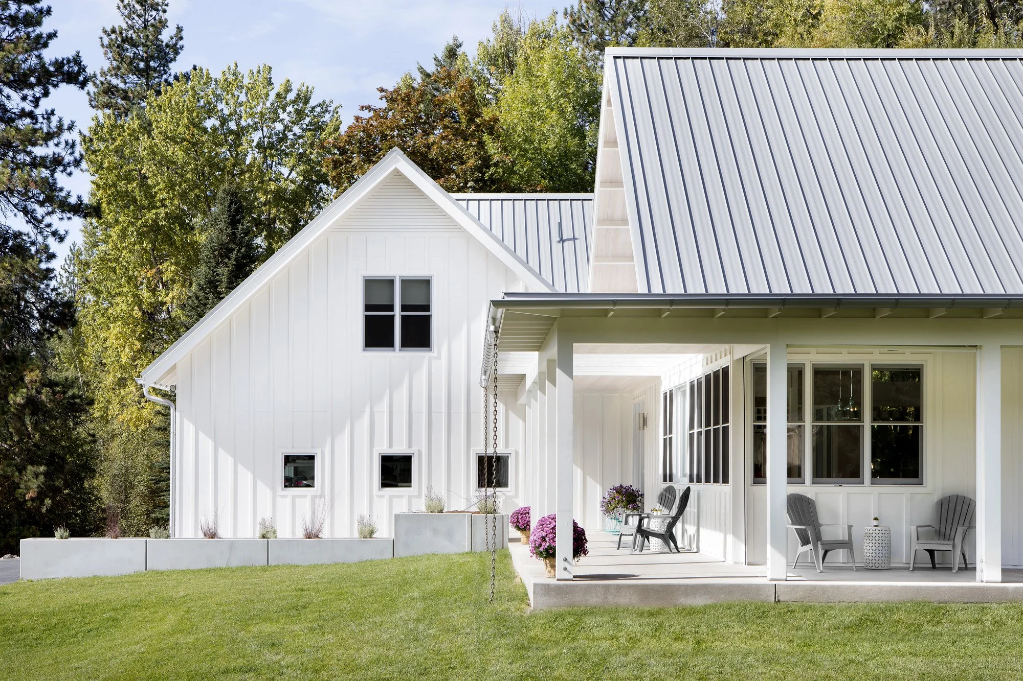 A modern white house with a metal roof and a spacious front porch with seating, surrounded by green grass and trees. Custom Home built in Montana by Edgell Building.