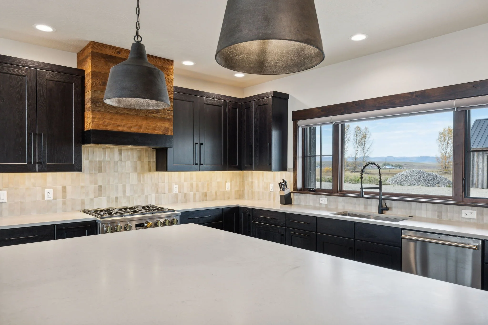 Modern kitchen with black cabinets, a white countertop, a large window showing a scenic countryside view, and two black pendant lights. Custom Home built in Montana by Edgell Building.