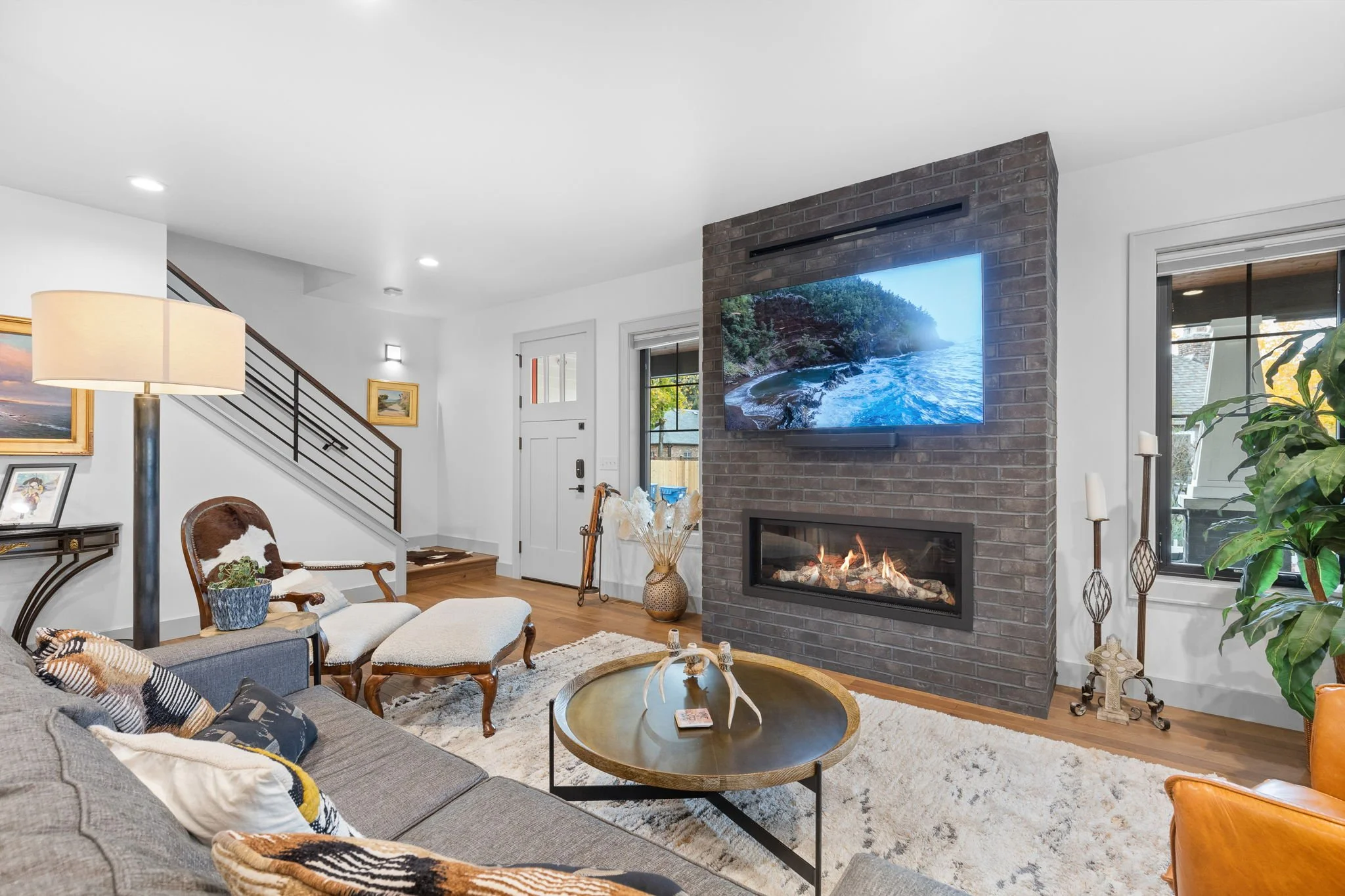 Living room with a gray sofa, cowhide armchair, and a circular wooden coffee table, a brick wall with a mounted TV and fireplace, and various plants and decorative items. Custom Home built in Montana by Edgell Building.