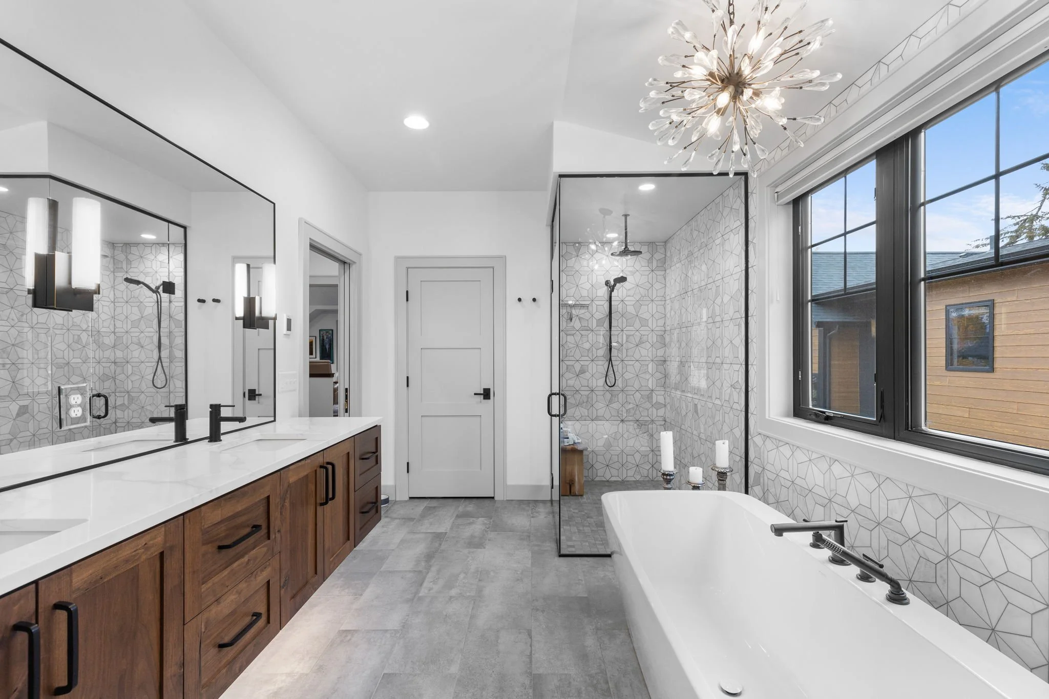 Modern bathroom with a double vanity, white countertop, black fixtures, large mirror, a walk-in shower, and a bathtub with candles, large window, and a stylish chandelier. Custom Home built in Montana by Edgell Building.