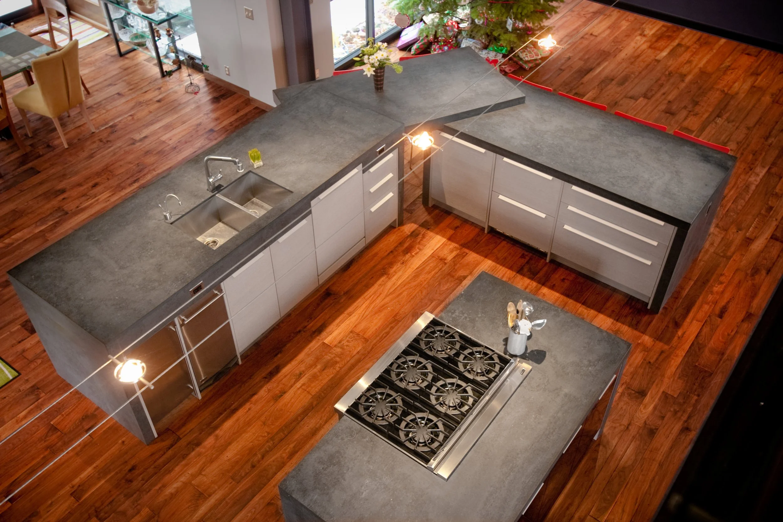 A modern kitchen with dark gray countertops, white cabinets, a stainless steel stove, a double sink, and hardwood floors. A Christmas tree is decorated in the background near a sliding glass door. Custom Home built in Montana by Edgell Building.
