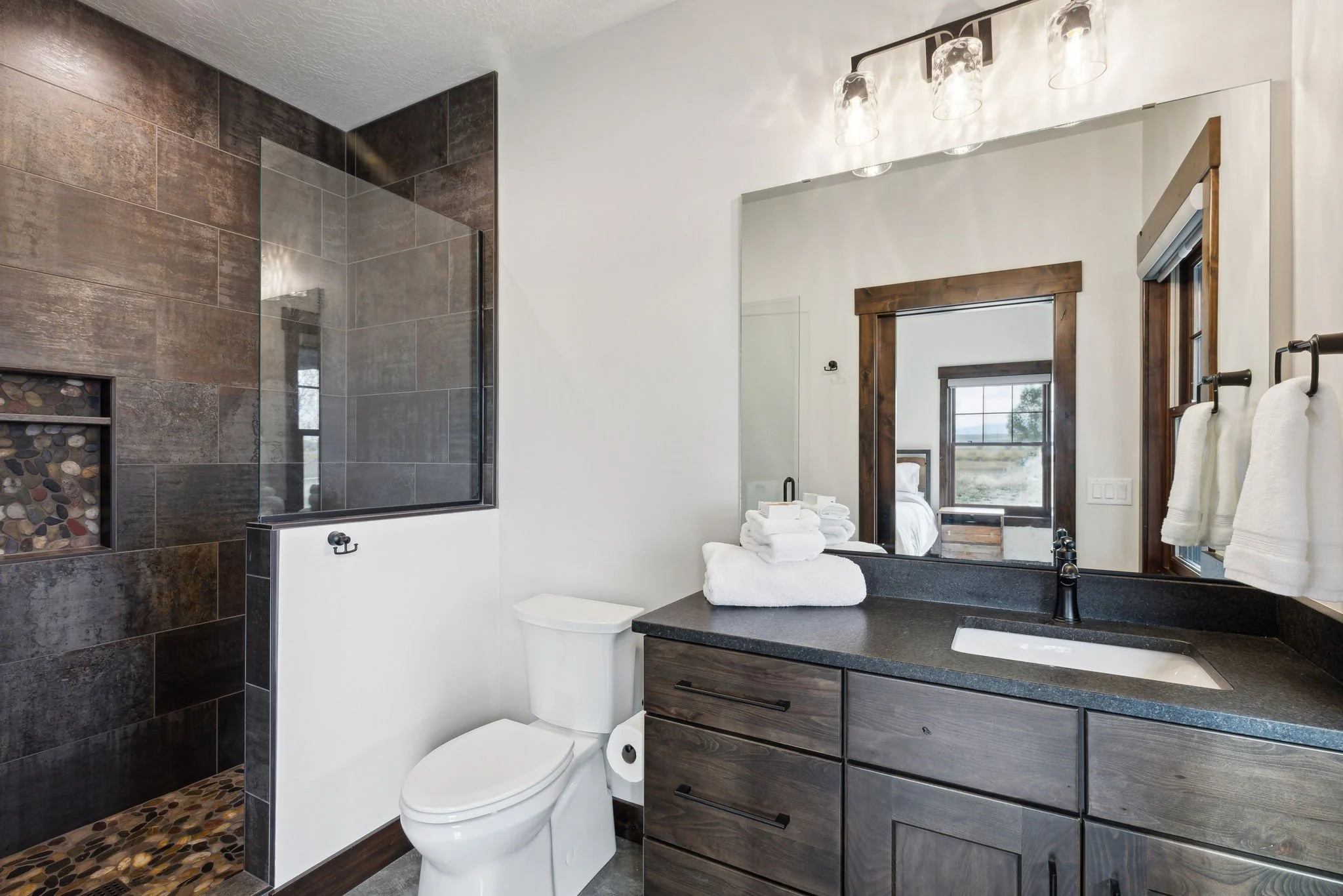 Modern bathroom with a walk-in shower featuring dark tiles and pebble floor, a toilet, a wooden vanity with a black countertop, a mirror, and towel racks with white towels. Custom Home built in Montana by Edgell Building.