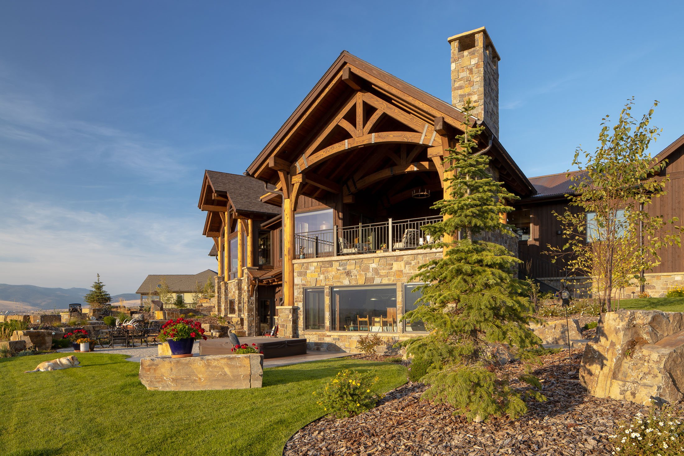 A large house with wooden and stone exterior, a chimney, a covered balcony, and a landscaped yard with green grass, potted flowers, and a dog lying on the lawn. Custom Home built in Montana by Edgell Building.