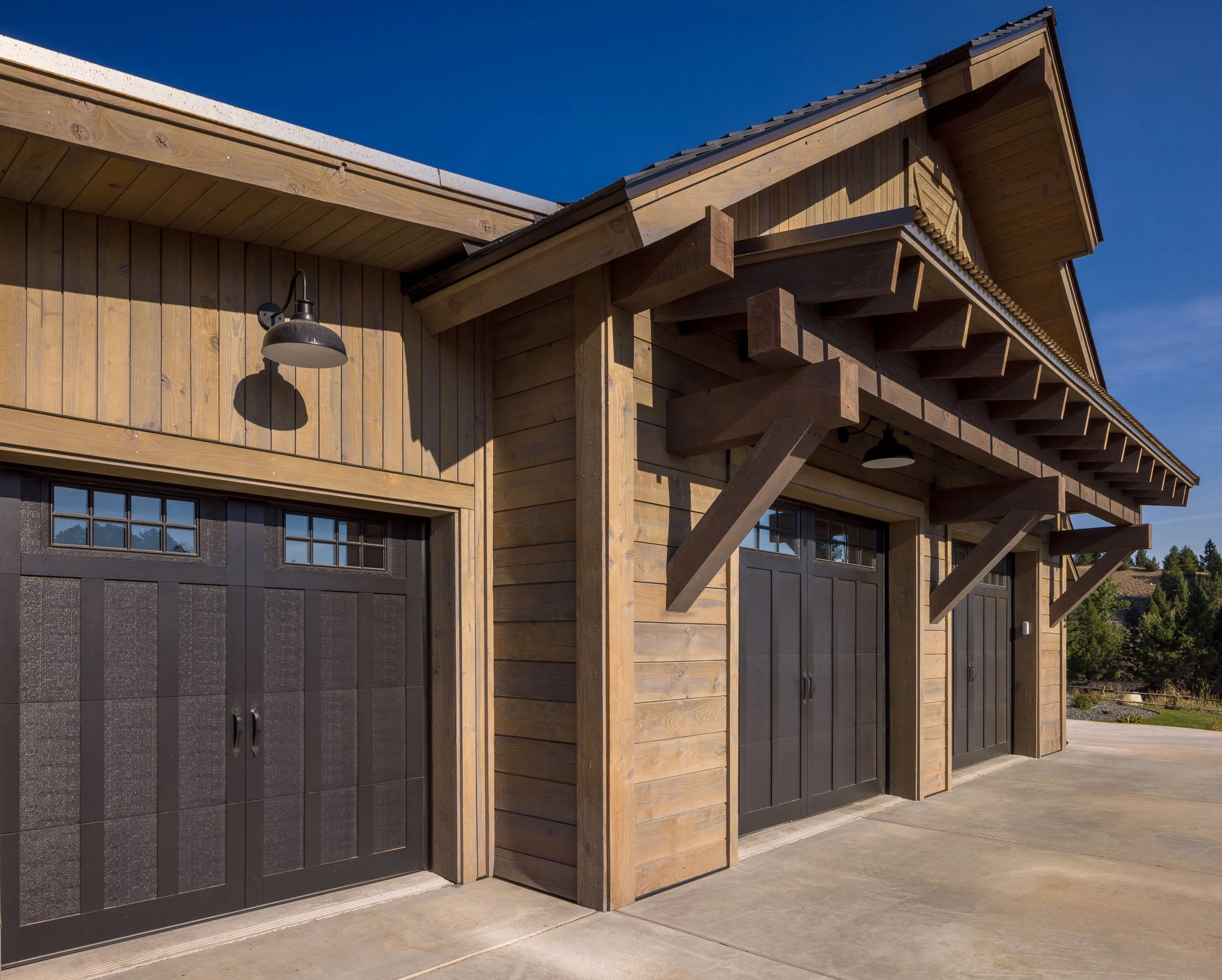 Three black garage doors with small windows at the top, surrounded by wooden siding on a modern building, under a clear blue sky. Custom Home built in Montana by Edgell Building.