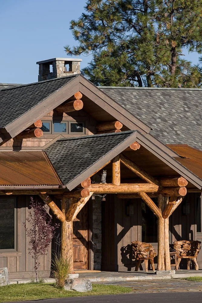 Close-up of a rustic log cabin entrance with wooden beams, log accents, and outdoor chairs, surrounded by trees. Custom Home built in Montana by Edgell Building.
