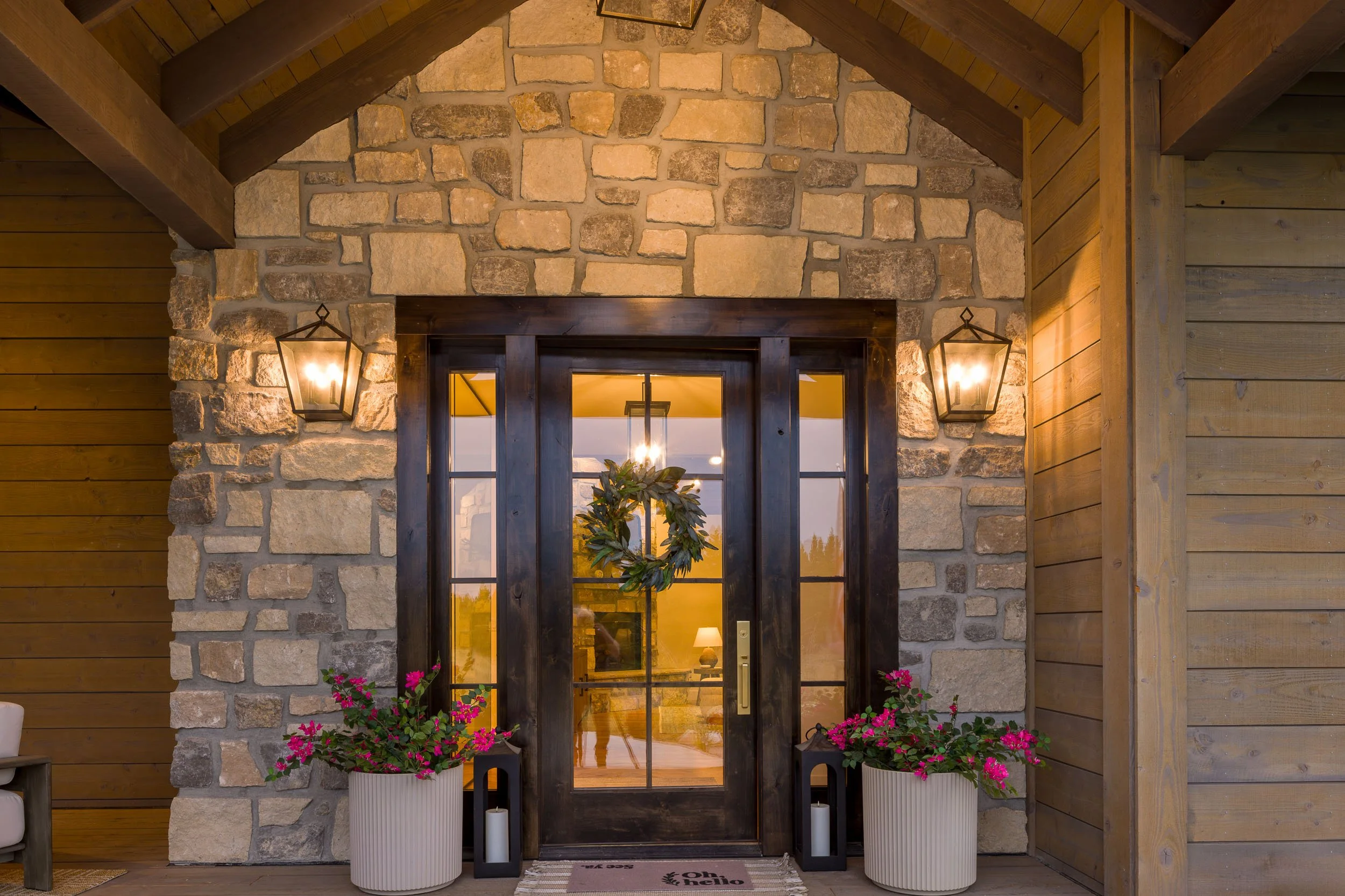 Front porch entrance with a wooden door, stone and wood siding, outdoor lanterns, potted flowers, and a wreath on the door. Custom Home built in Montana by Edgell Building.