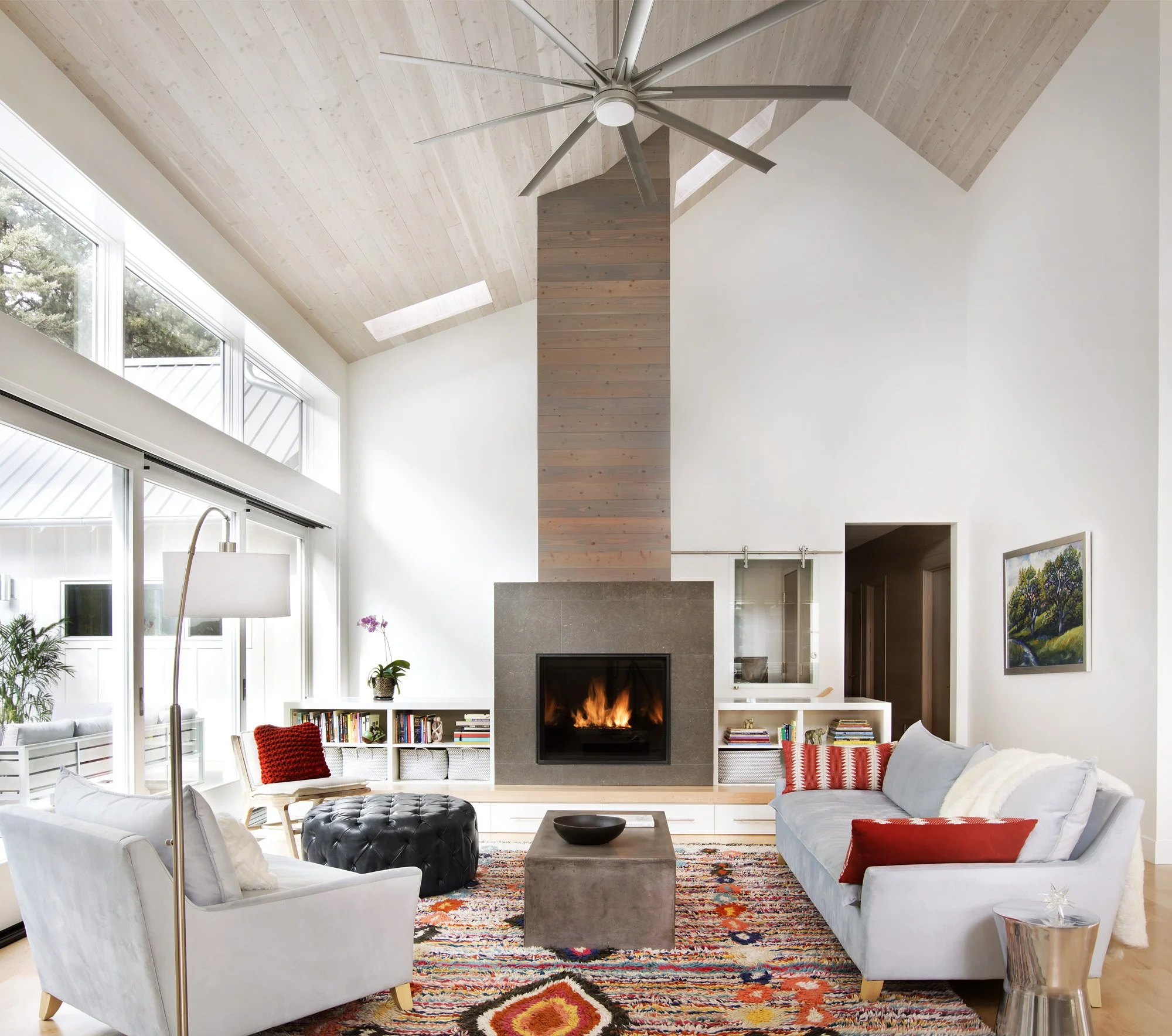 Living room with a fireplace, two white sofas, colorful rug, and tall windows with a view of trees outside. Custom Home built in Montana by Edgell Building.