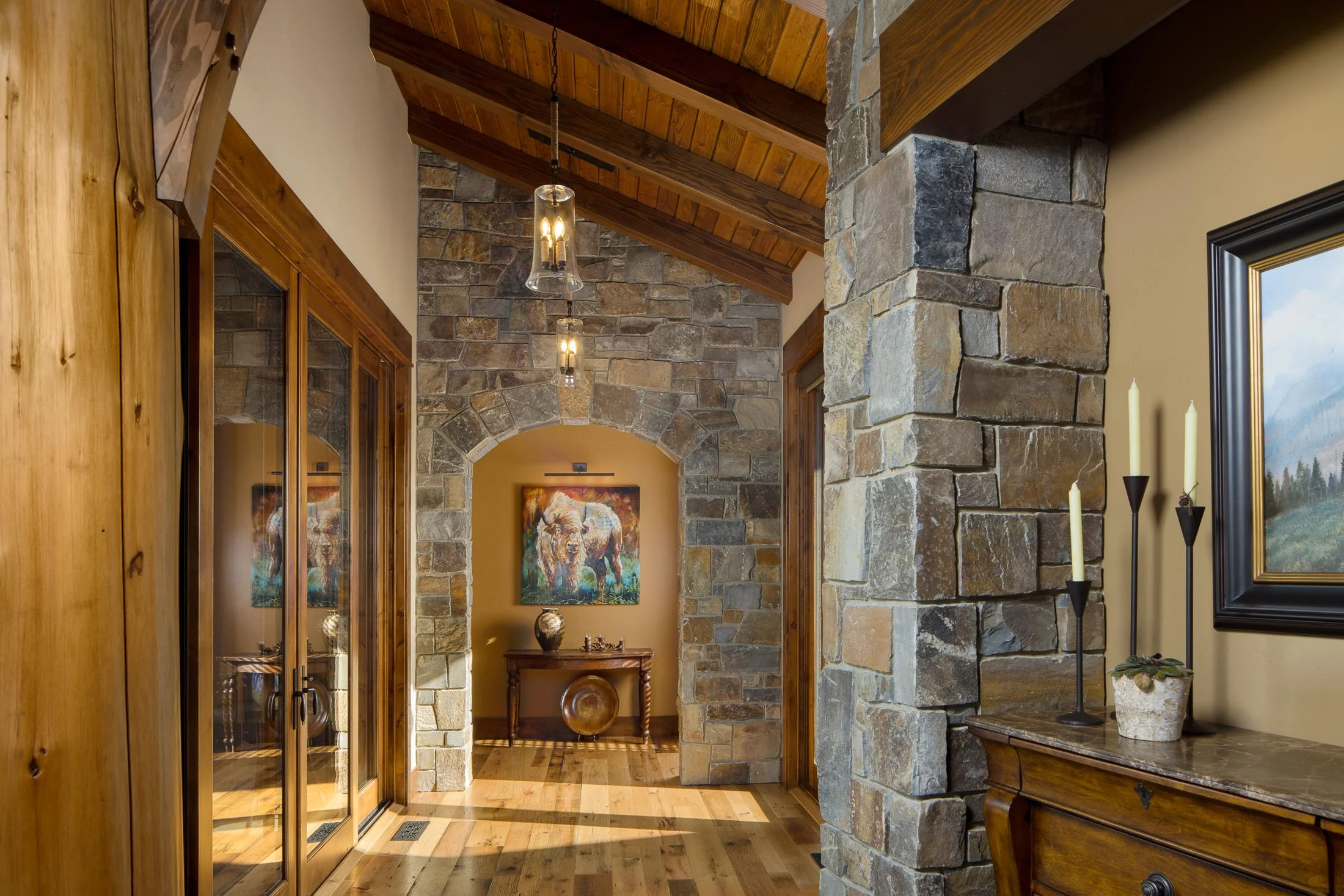 A hallway with wooden and stone walls, wooden ceiling with pendant lights, featuring an arched stone doorway leading to a small table with decorative vases and a painting of a buffalo on the wall. Custom Home built in Montana by Edgell Building.
