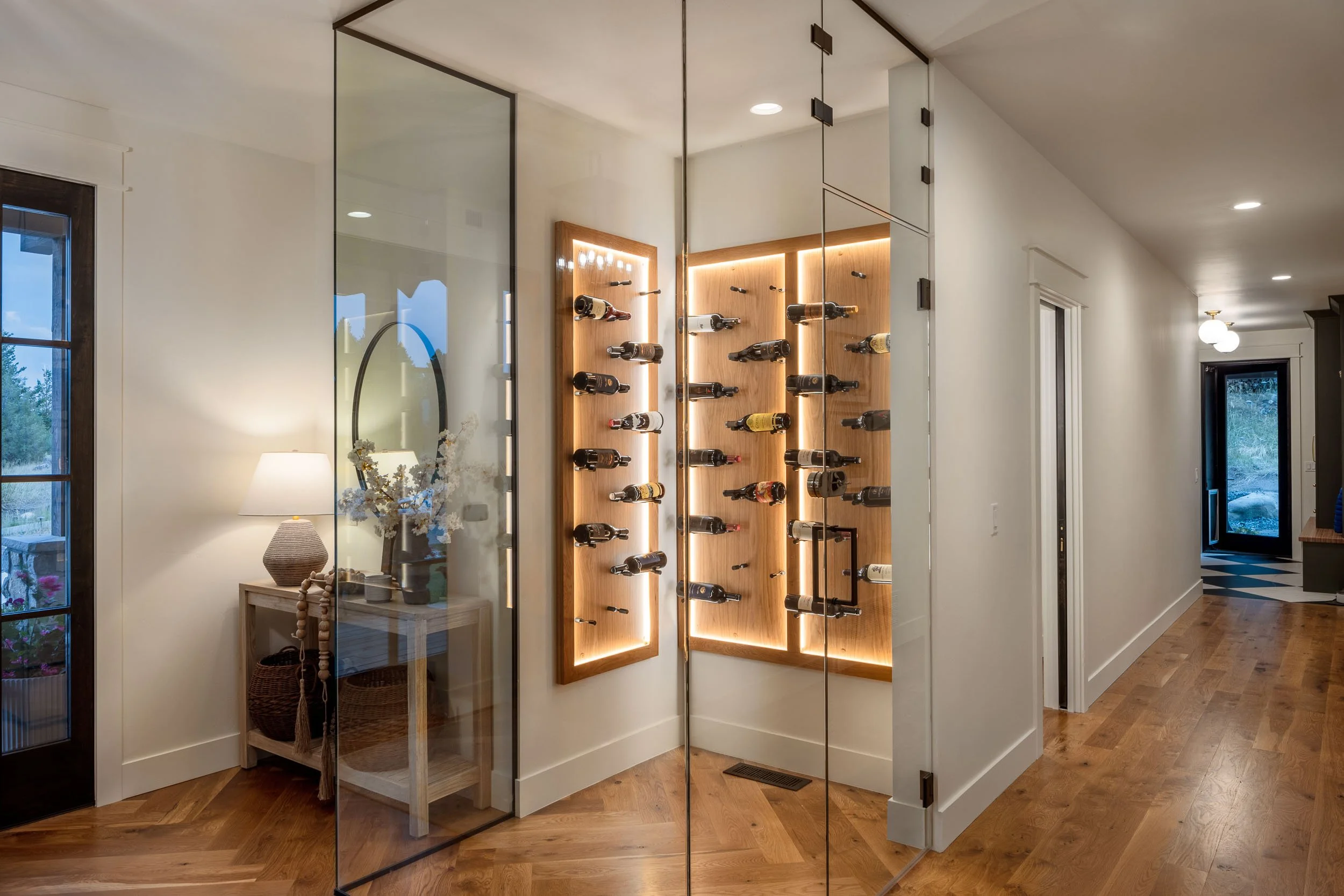 Interior of a home showing a glass-walled wine storage area with wooden shelves and bottles, a side table with a lamp and decorative items. Home Designed and Built By Edgell Building in Western Montana.