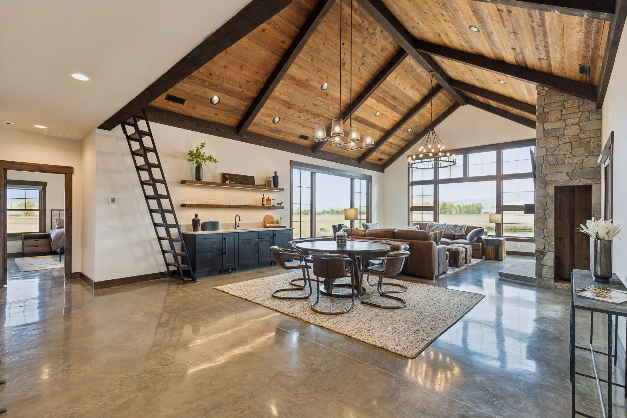 Open-plan living room with high vaulted wooden ceiling, large windows, stone fireplace, and modern furniture. Custom Home built in Montana by Edgell Building.