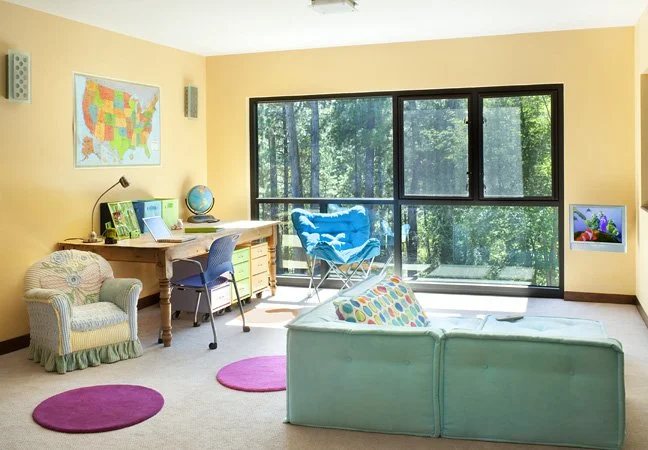 Brightly lit classroom with yellow walls, a large window with a view of trees, a desk with a globe, a colorful chair, a small armchair, a light green sofa with a polka dot pillow, and three pink rugs.  Custom Home built in Montana by Edgell Building.