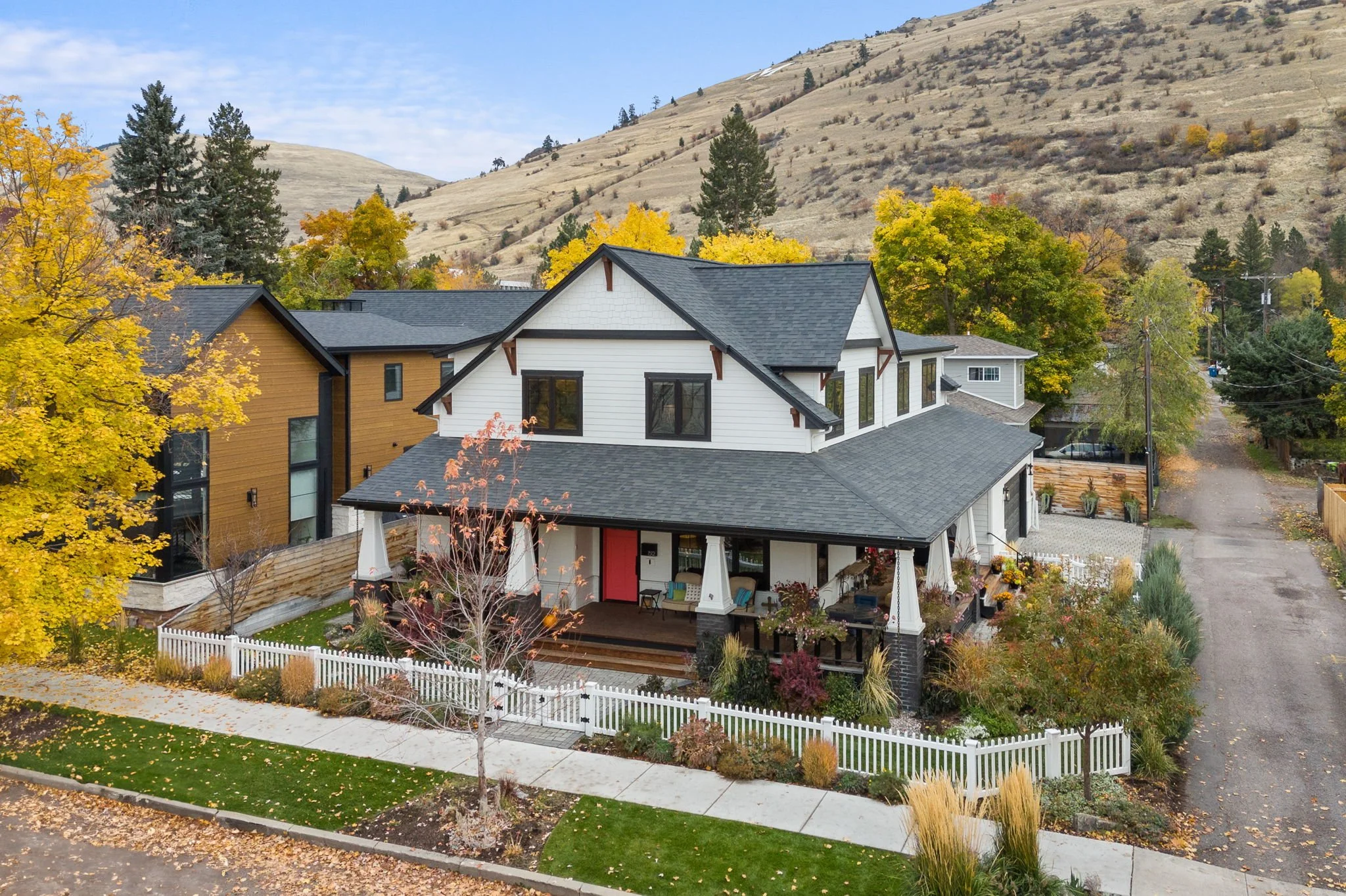 A two-story house with a porch, black trim, and a pink front door, surrounded by a white picket fence, lawn, and fall foliage, with a hillside in the background. Custom Home built in Montana by Edgell Building.