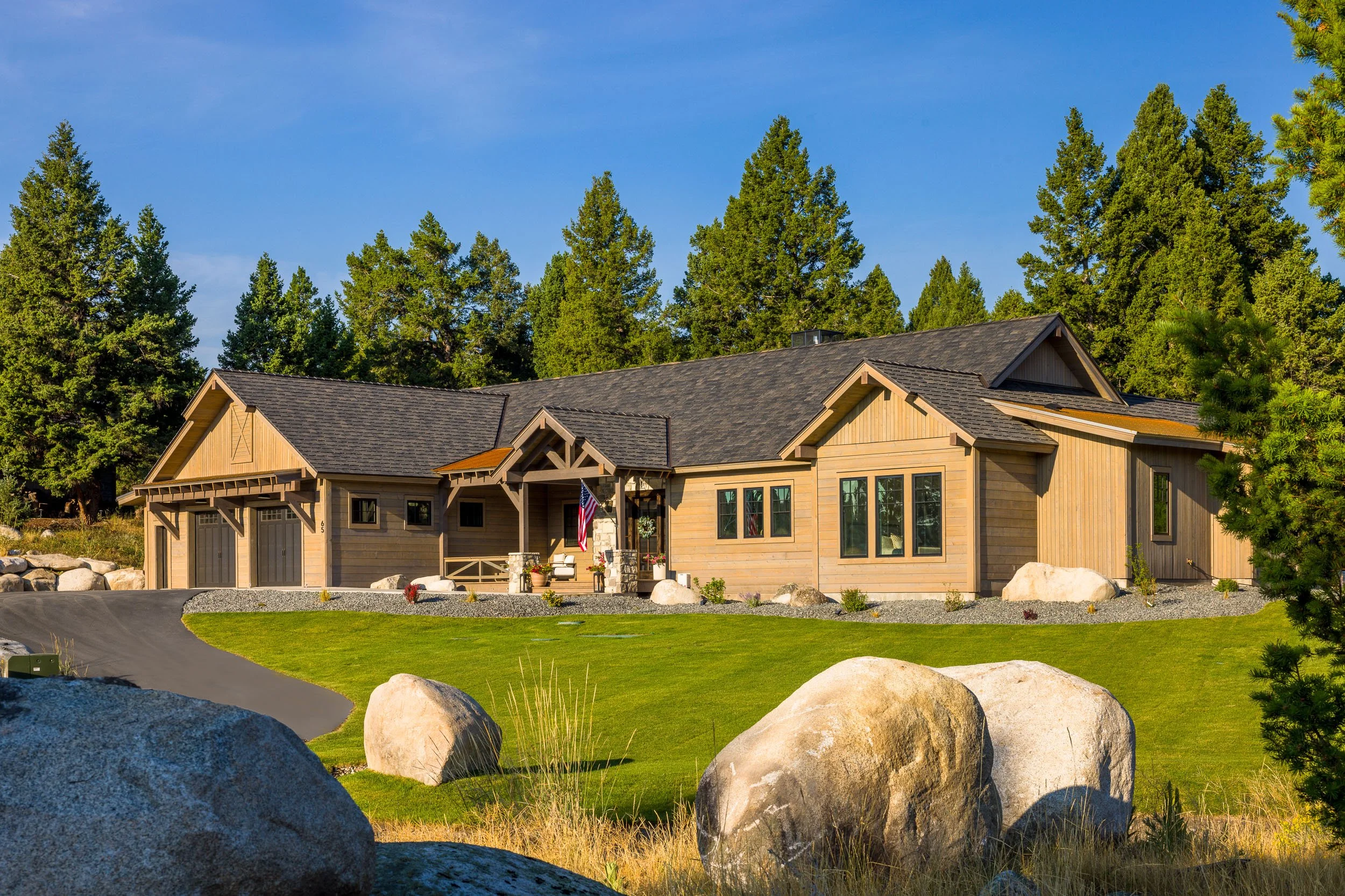 A modern house with a wooden exterior, large windows, a two-car garage, and landscaped front yard with grass, rocks, and trees under a clear blue sky. Custom Home built in Montana by Edgell Building.