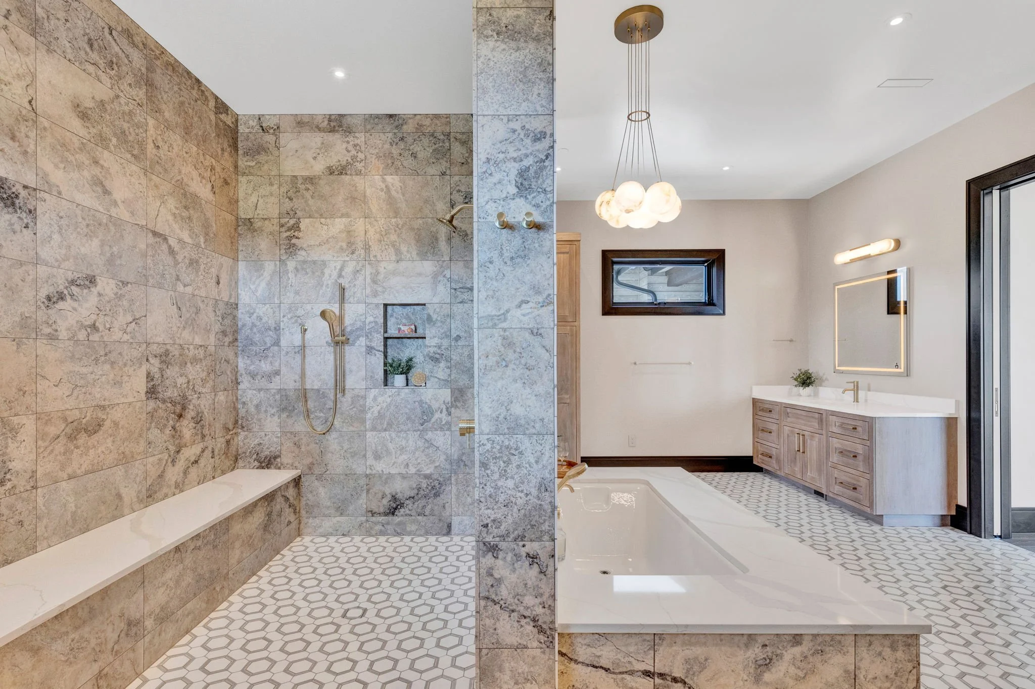 A bathroom with a separate walk-in shower and a large bathtub, featuring beige and gray tiles, a vanity with a mirror and lighting, and a window with dark trim. Custom Home built in Montana by Edgell Building.
