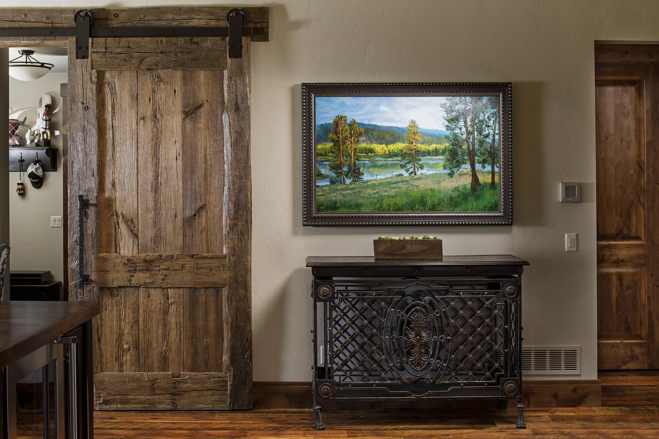 Interior scene with a rustic wooden sliding barn door, a landscape painting of trees and a river on the wall, a decorative black metal table below the painting, and hardwood flooring. Custom Home built in Montana by Edgell Building.
