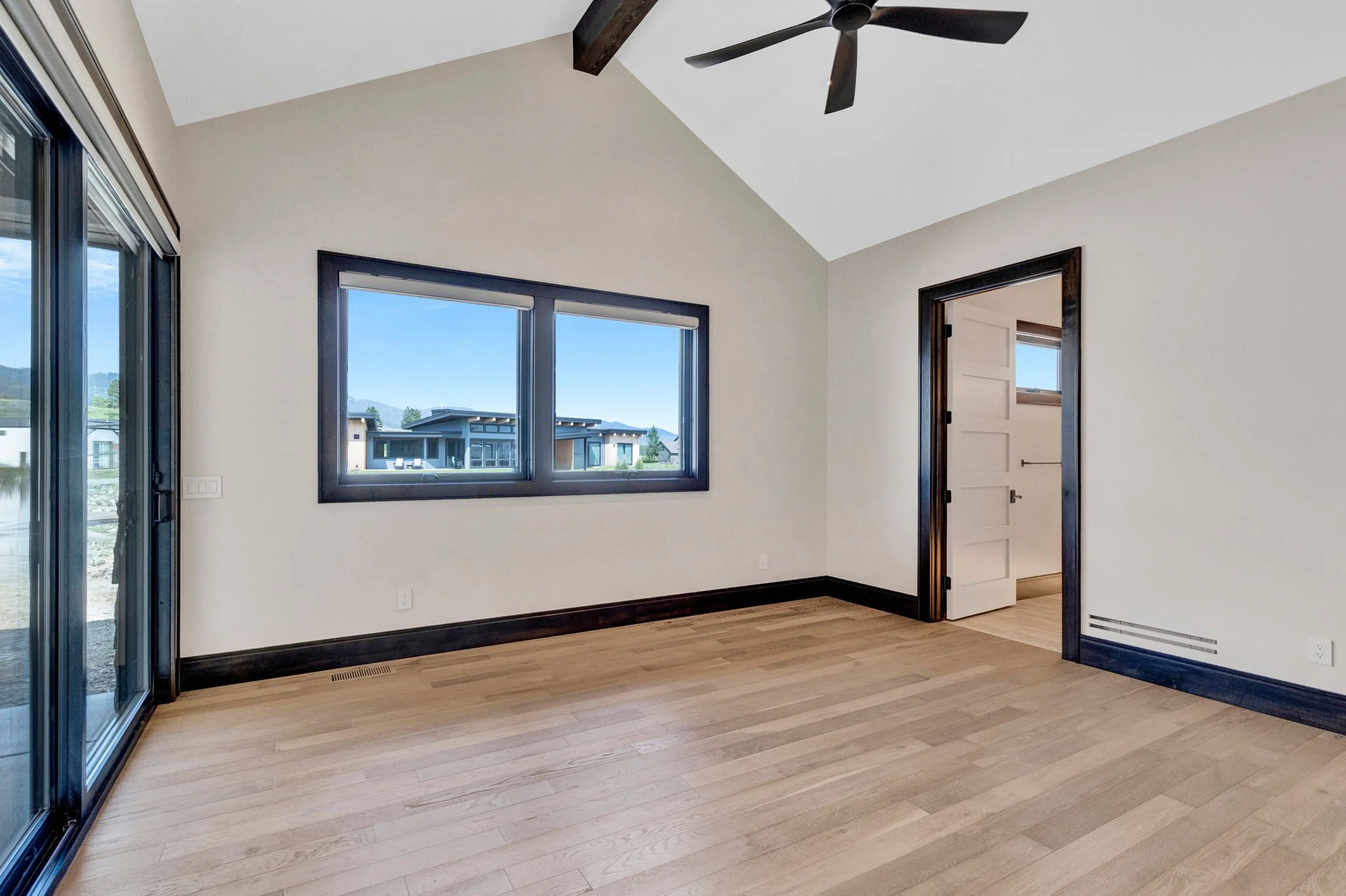Empty room with light hardwood floors, beige walls, large windows, and a ceiling fan. Custom Home built in Montana by Edgell Building.