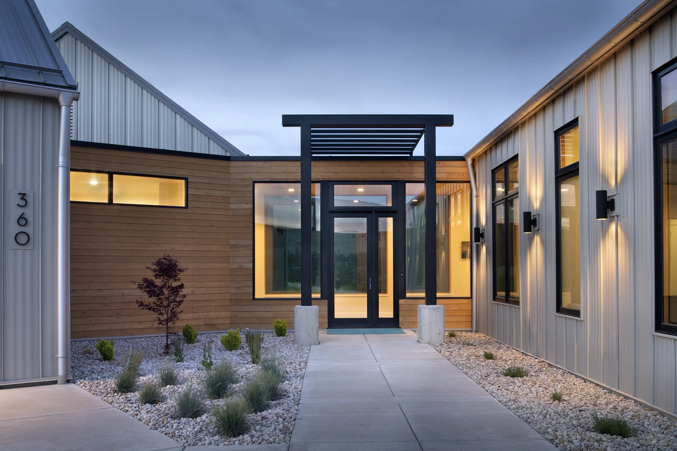 Modern residential building with wood and metal siding, illuminated windows, landscaped front yard with small plants and gravel, concrete pathway, and a black pergola structure at the entrance. Custom Home built in Montana by Edgell Building.