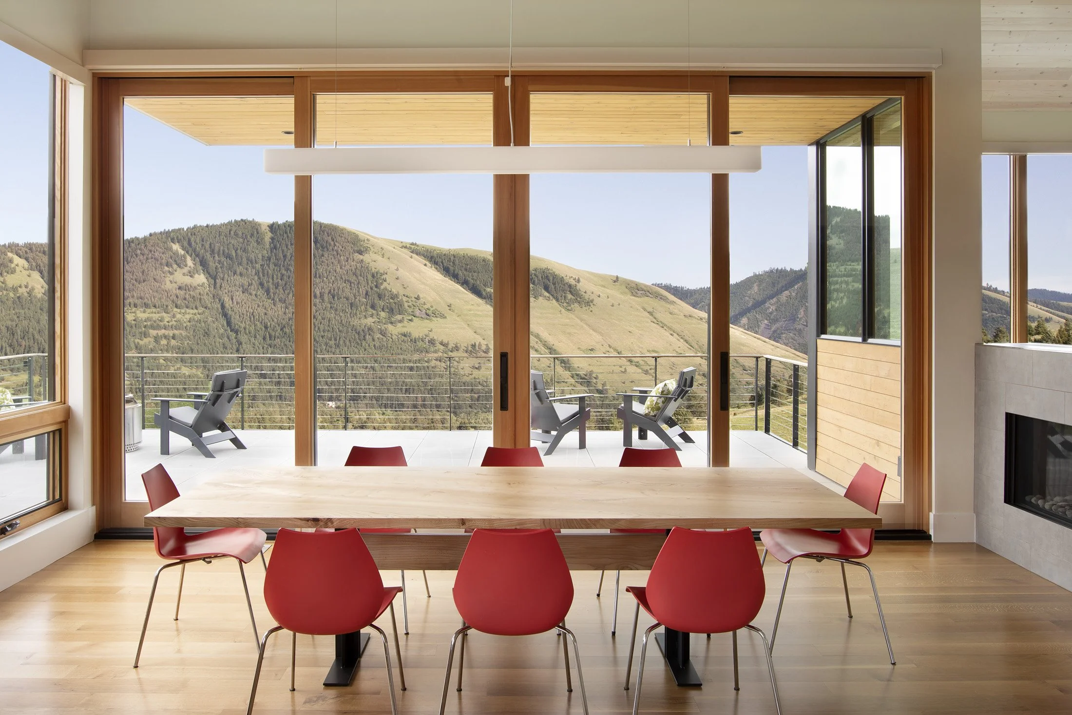 Modern dining room with large sliding glass doors opening to a mountain view, featuring a wooden dining table and six red chairs. Outside, there is a balcony with two chairs and a mountain landscape.  Custom Home built in Montana by Edgell Building.