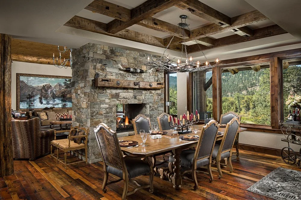 A rustic dining room with a large wooden table, eight chairs, and a stone fireplace with a fire burning. The room has large windows showing a green mountain landscape, hardwood floors.  Custom Home built in Montana by Edgell Building.