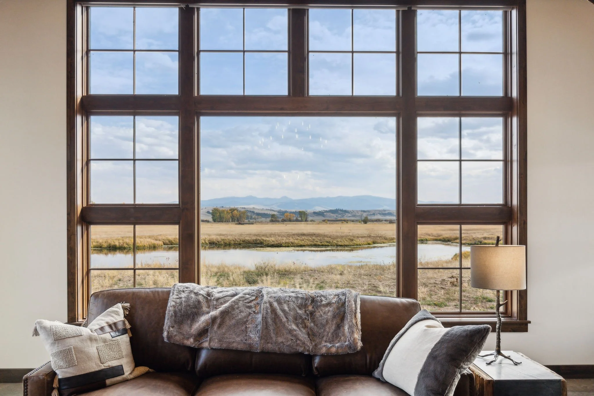 Living room with a large window overlooking a field, river, and mountains, with a brown sofa, pillows, a fur throw, and a floor lamp. Custom Home built in Montana by Edgell Building.