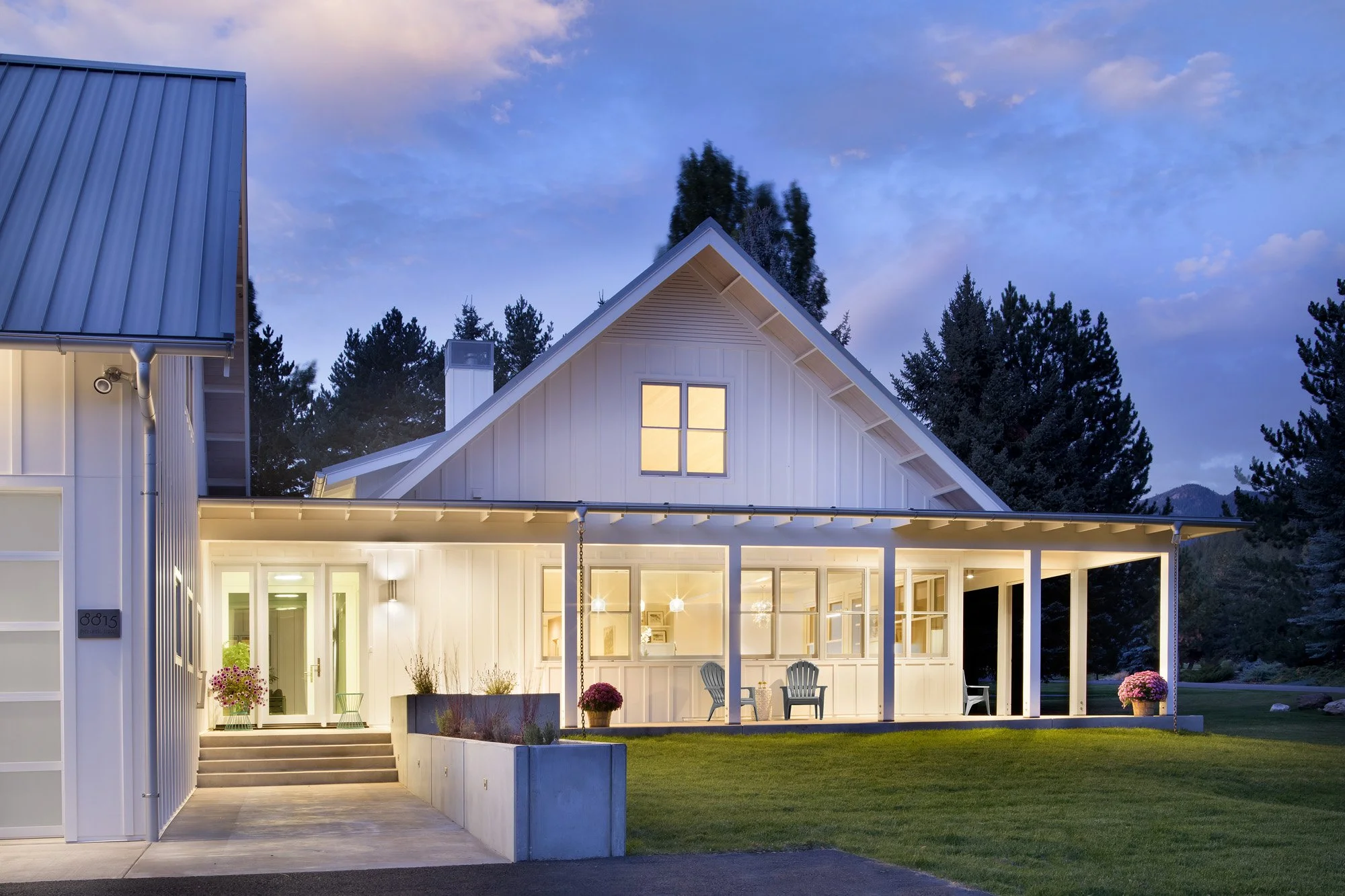 A modern white house with a sloped roof and large windows illuminated at dusk, surrounded by a green lawn and tall trees in the background. Custom Home built in Montana by Edgell Building.