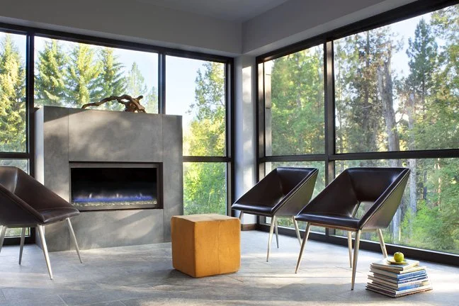 Modern living room with large windows showing trees outside, two black armchairs, a small orange ottoman, a stack of books, and a fireplace with a piece of driftwood on top. Custom Home built in Montana by Edgell Building.