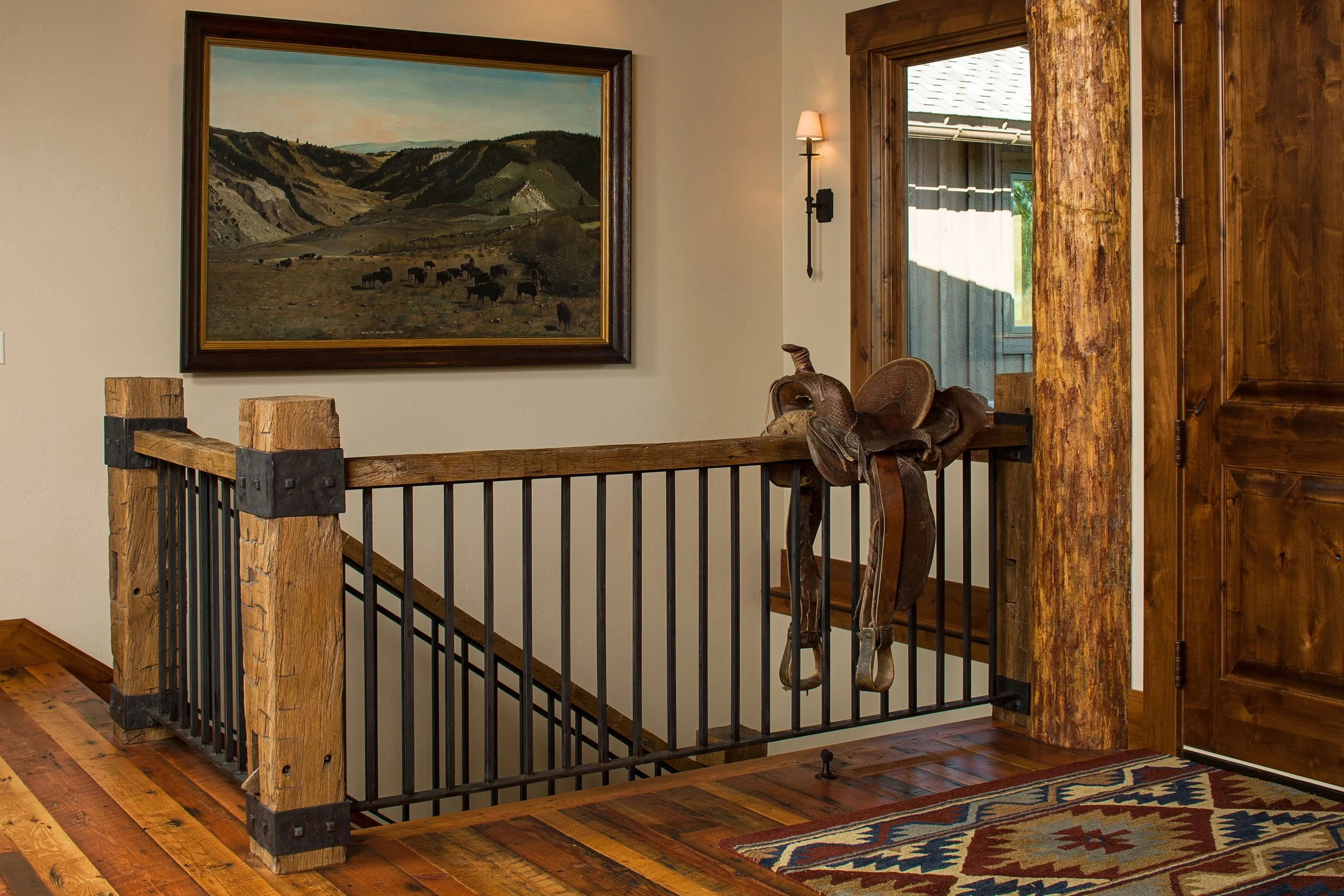 Interior of a rustic house with wooden staircase and railing, a painting of mountains and cattle on the wall, and a window with a door nearby, decorated with a saddle hanging on the railing. Custom Home built in Montana by Edgell Building.