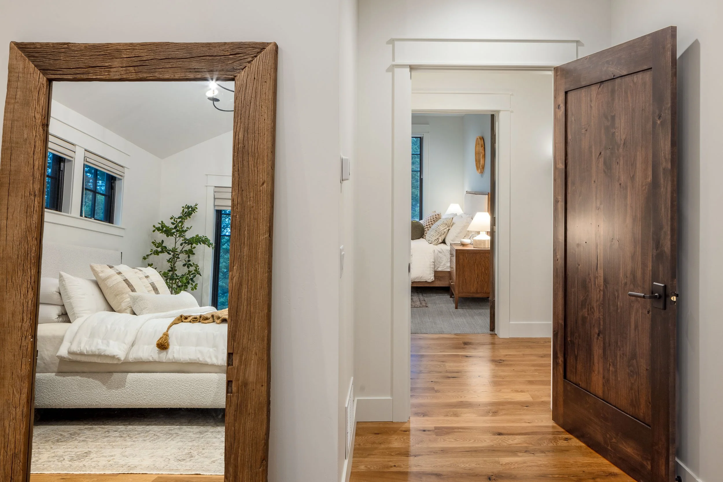 View from a hallway into a bedroom, with a large rustic wooden framed mirror on the left reflecting the room, and another open wooden door on the right revealing the bedroom with a bed, lamps, and windows.