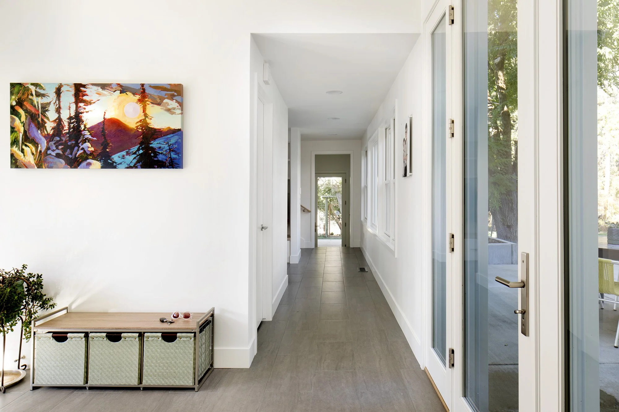 Bright hallway with white walls, large windows, and door leading outside, with artwork on wall and a small storage bench with plants nearby. Custom Home built in Montana by Edgell Building.