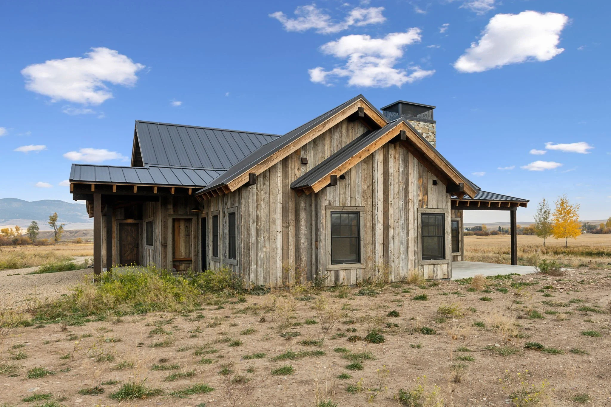 A rustic wooden house with a metal roof in a rural landscape under a blue sky with scattered clouds. Custom Home built in Montana by Edgell Building.