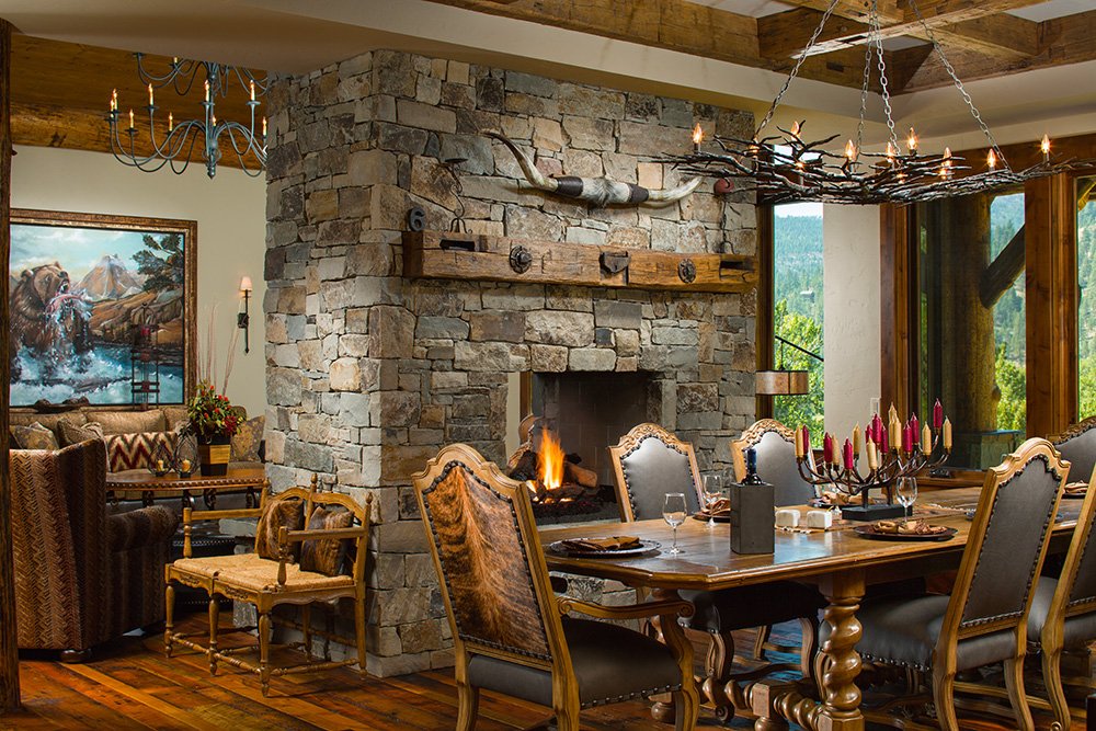 A rustic dining room with a stone fireplace, a wooden chandelier, and a large wooden dining table set with candles and glassware. There are large windows with a view of greenery outside. Custom Home built in Montana by Edgell Building.