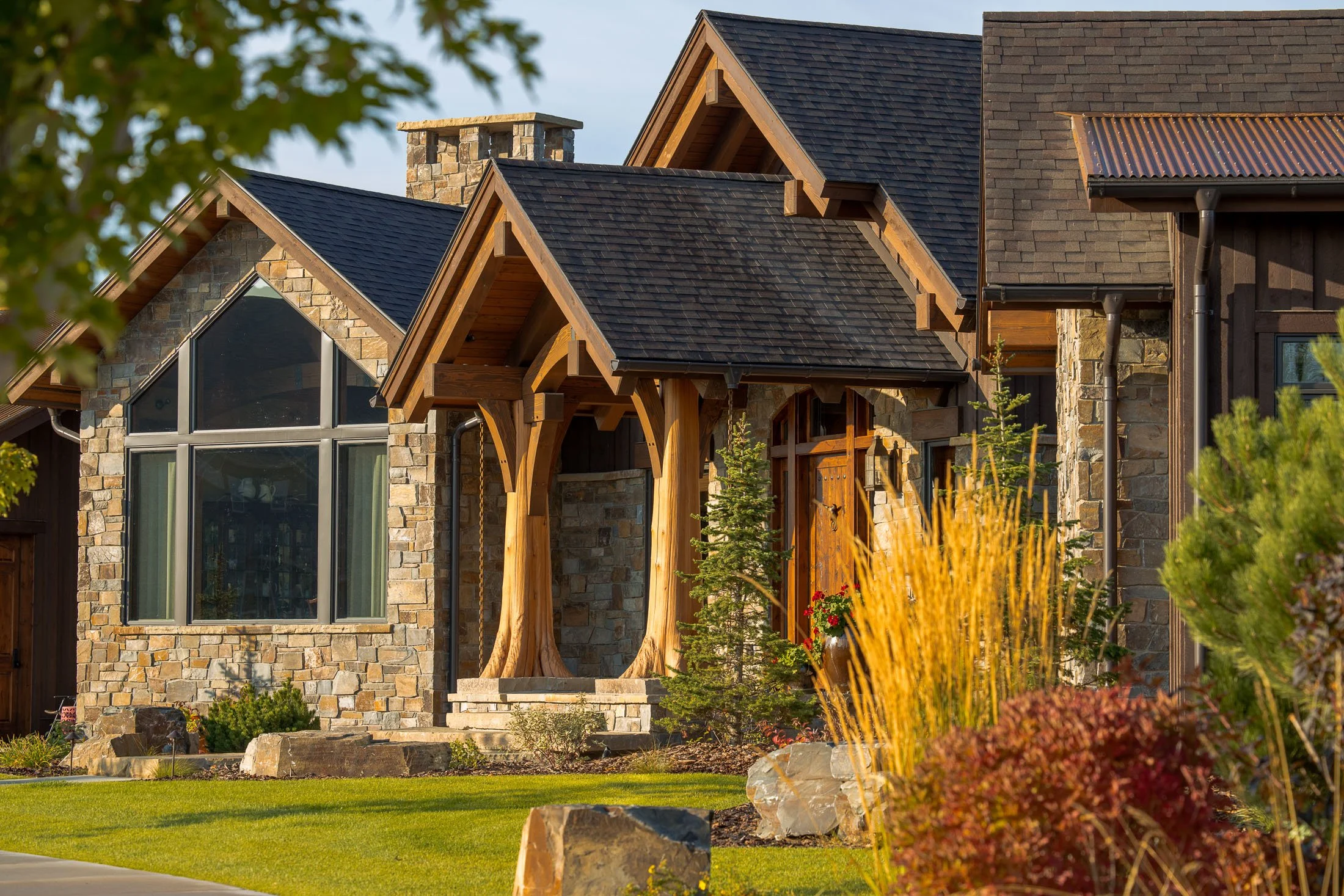 Front exterior of a modern house with stone and wood siding, large windows, and a covered porch supported by wooden beams, with landscaped front yard and plants. Custom Home built in Montana by Edgell Building.