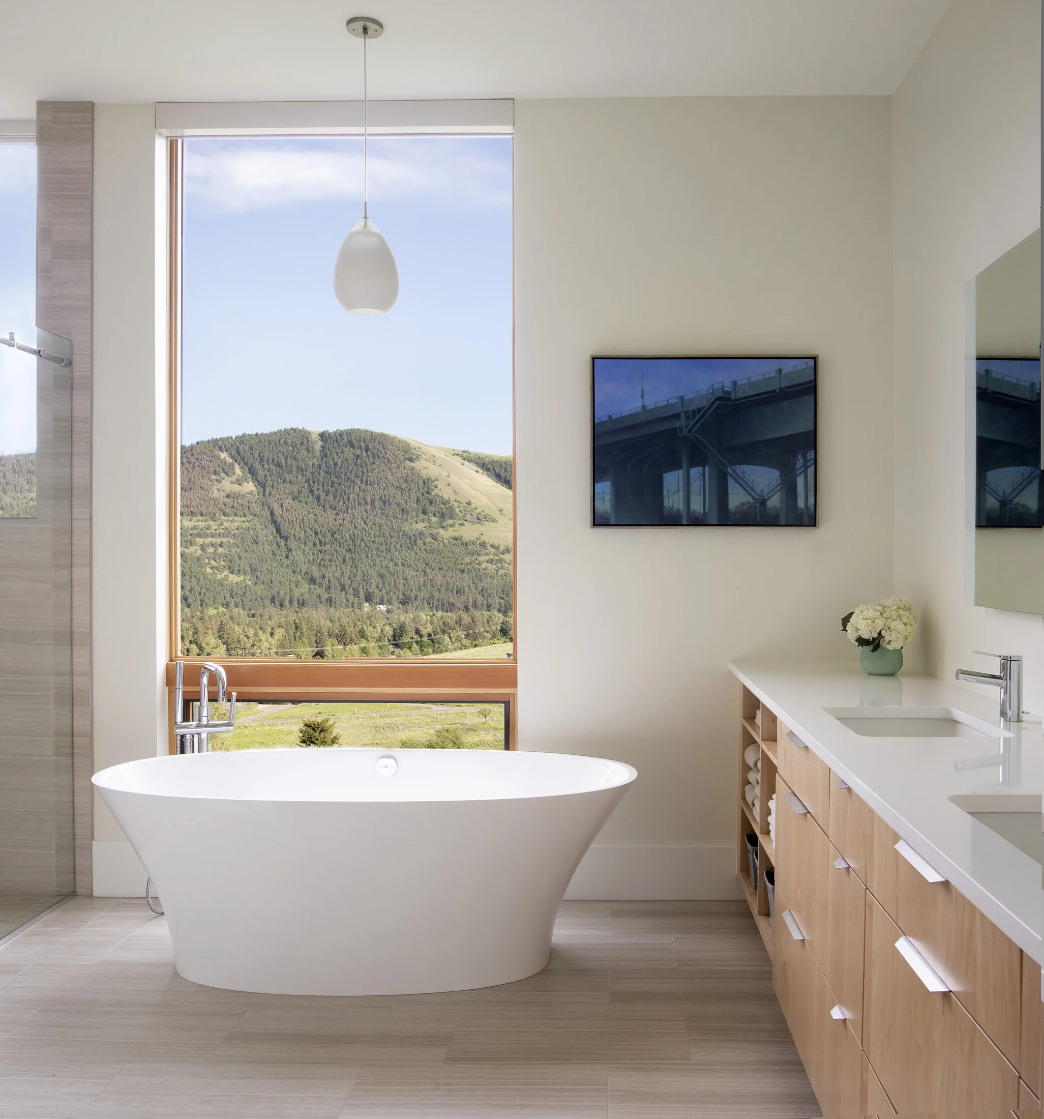 Modern bathroom featuring a freestanding bathtub by a large window with a mountain view, with a wooden vanity, a mounted TV, and a small bouquet of white flowers. Custom Home built in Montana by Edgell Building.