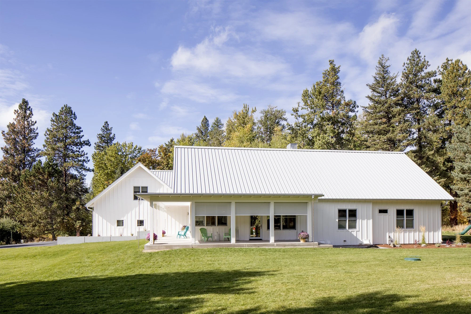 A white house with a metal roof surrounded by trees, with a green lawn in front and a porch with outdoor chairs. Custom Home built in Montana by Edgell Building.