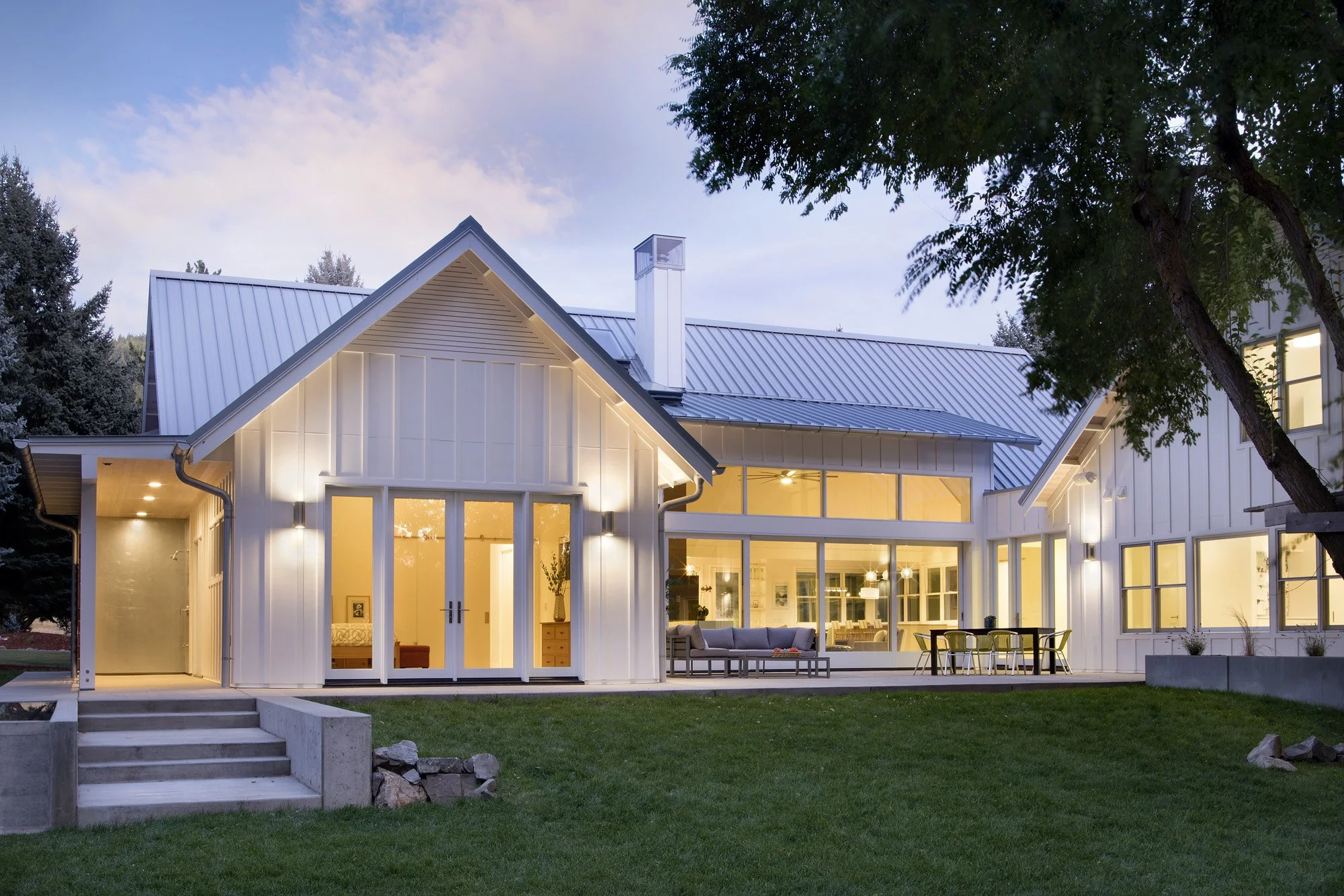 Modern house with white exterior walls and a metal roof, lit from inside, surrounded by a green lawn and trees, taken at dusk. Custom Home built in Montana by Edgell Building.