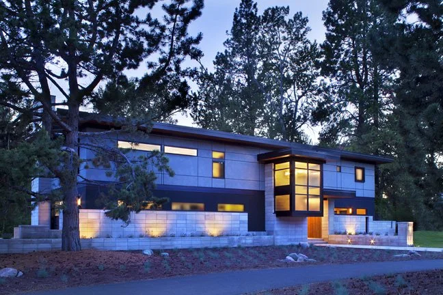 Modern two-story house with large windows, situated among tall trees, illuminated at dusk. Custom Home built in Montana by Edgell Building.
