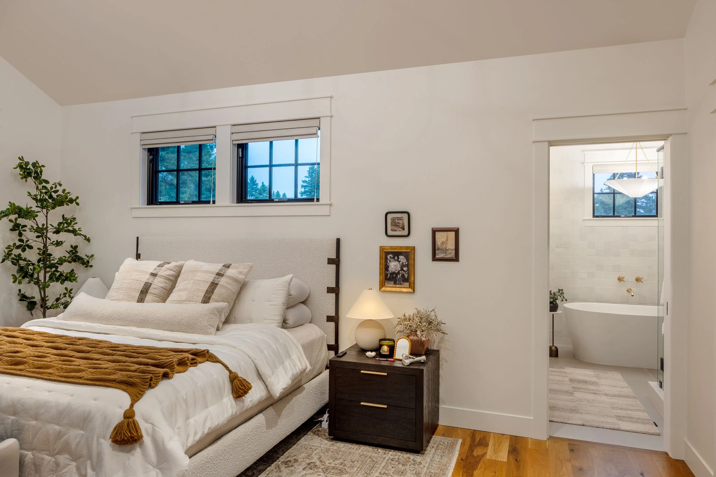 A bedroom with a bed, pillows, nightstand with lamp, and bathroom with bathtub visible through an open doorway. Home Designed and Built By Edgell Building in Western Montana.