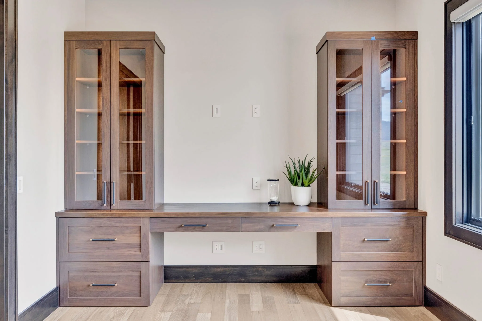 Empty wooden desk with glass display cabinets on each side, a potted plant, and an hourglass on top, set against a white wall near a window. Custom Home built in Montana by Edgell Building.