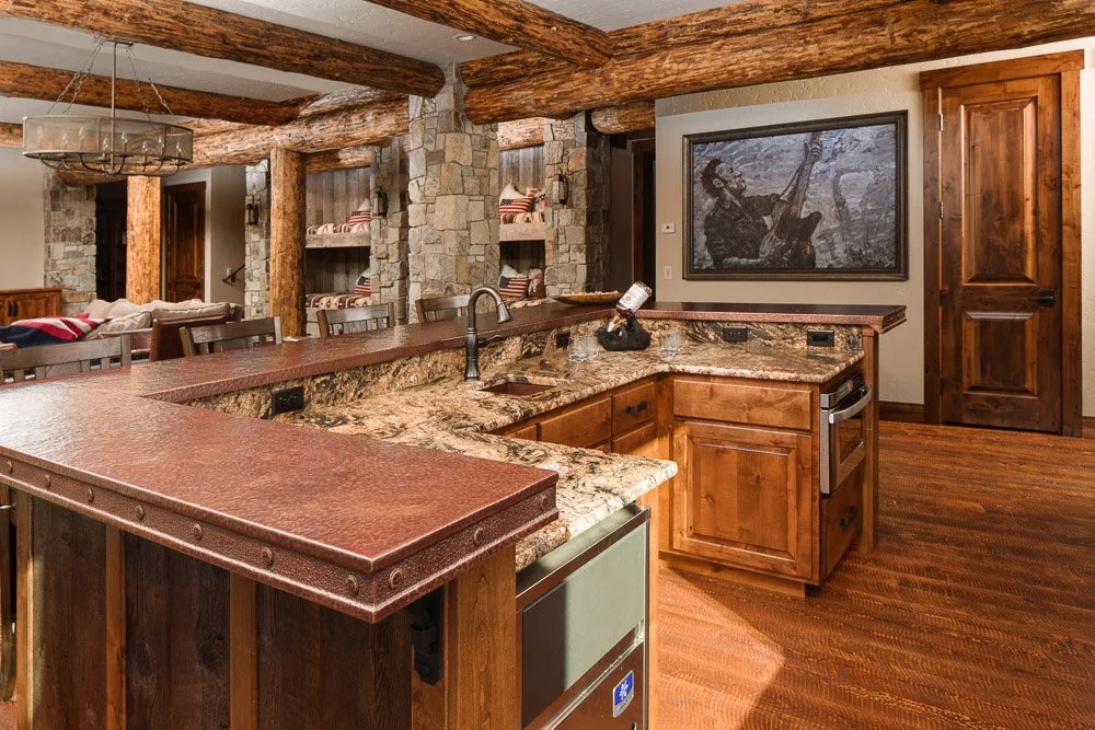 Rustic kitchen with wooden beams, stone walls, granite countertops, and a painted mural of a musician playing a guitar. Custom Home built in Montana by Edgell Building.